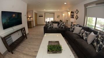 A living room with a large flat-screen TV, a sofa set, a coffee table with a plant, and a kitchen in the background.