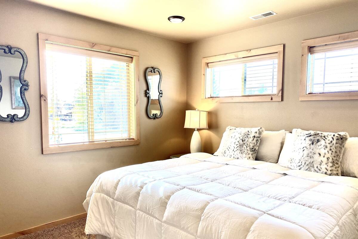 A bedroom with a large bed, two windows with blinds, a wall-mounted mirror, and a bedside lamp.