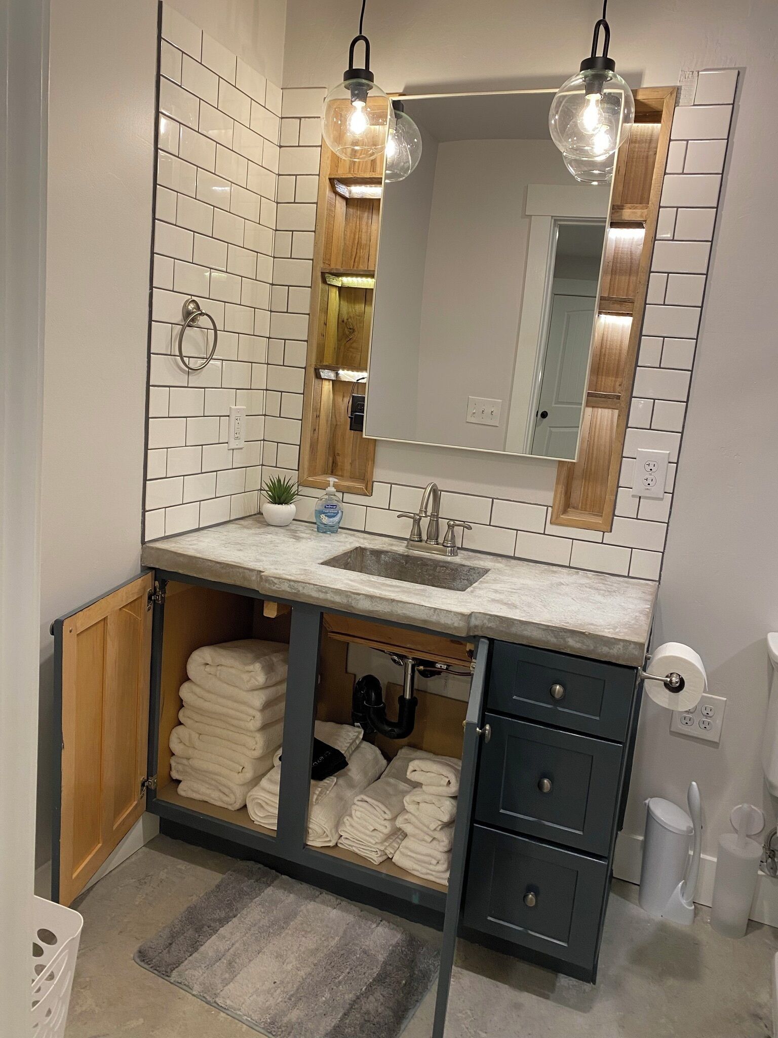 Une salle de bain moderne avec un mur recouvert de carreaux blancs, un meuble-lavabo en bois et un miroir au-dessus. Deux luminaires suspendus et un porte-serviettes sont fixés au mur.