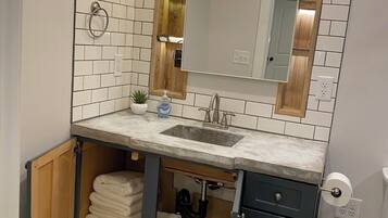 Une salle de bain moderne avec un mur recouvert de carreaux blancs, un meuble-lavabo en bois et un miroir au-dessus. Deux luminaires suspendus et un porte-serviettes sont fixés au mur.