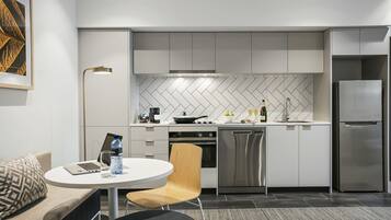 A modern kitchen with white cabinets, a herringbone backsplash, stainless steel appliances, and a dining area with a round table and a chair.