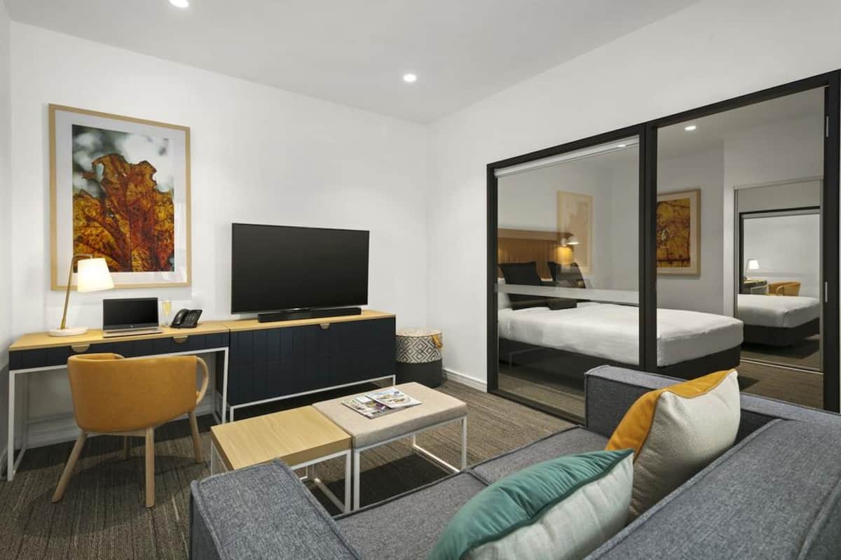 Habitación de hotel moderna con televisión de pantalla plana, escritorio con computadora portátil, sofá y cama con espejo grande.