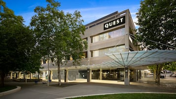 A modern building with a 'QUEST' sign, featuring a covered outdoor area with seating and a well-maintained lawn.
