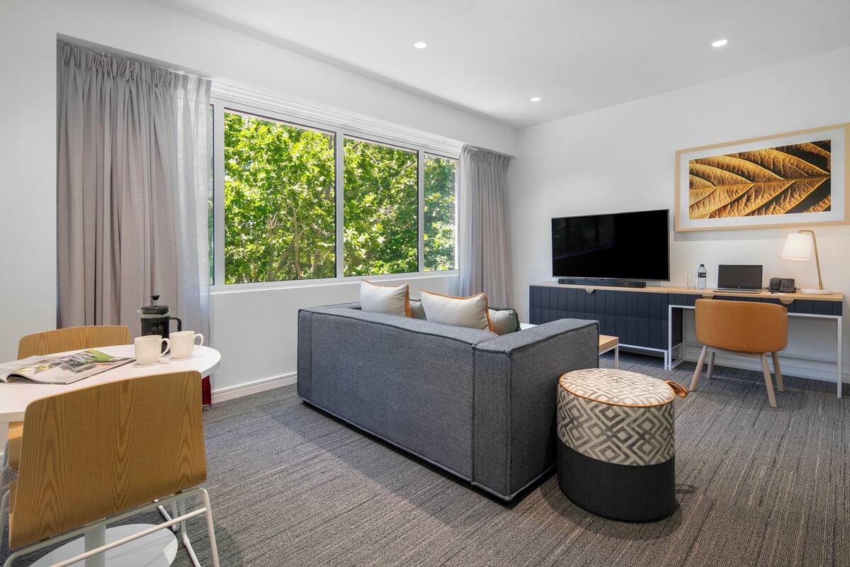 Una habitación de hotel moderna con un sofá gris, una mesa de comedor con sillas, un televisor y un escritorio con computadora.
