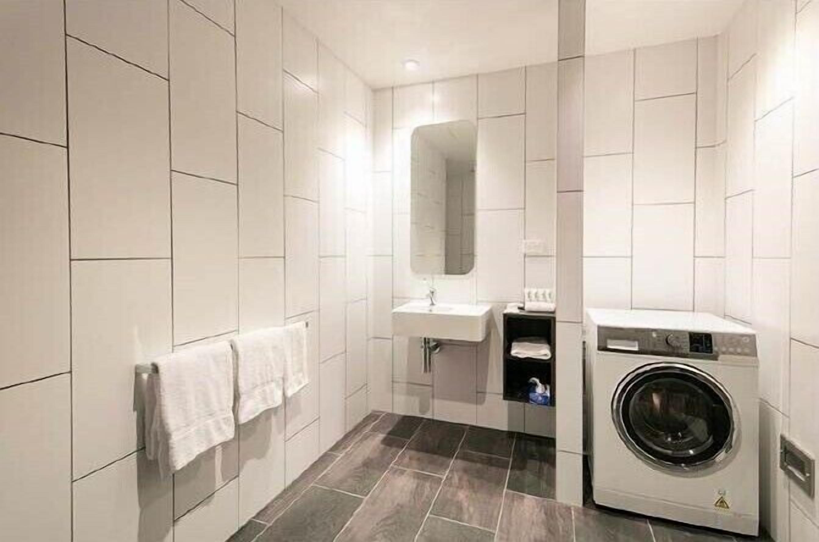 A modern bathroom with white tiles, a sink, a mirror, a towel rack, and a washing machine.