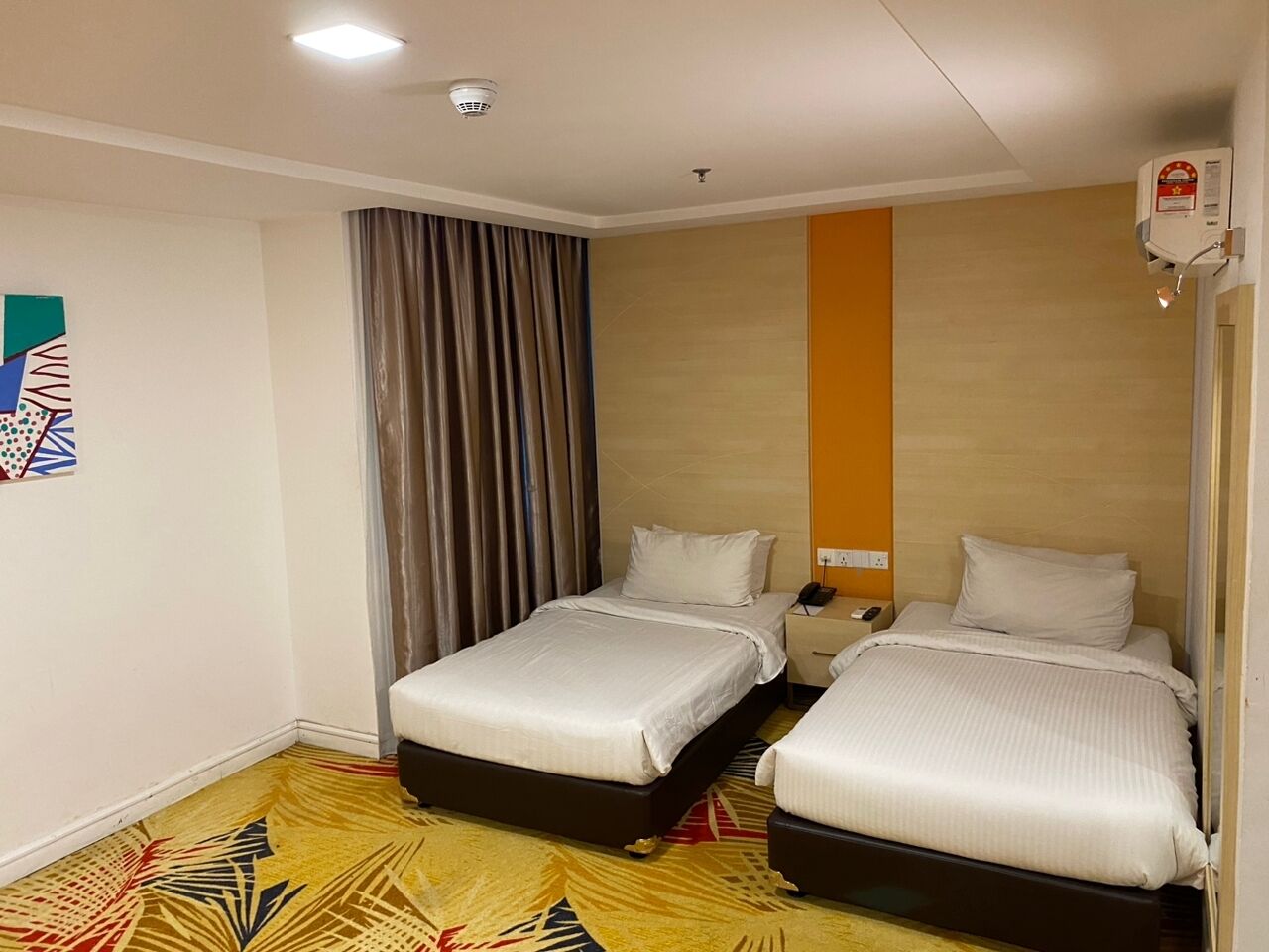 A hotel room with two beds, a large window with curtains, a wall-mounted air conditioning unit, and a patterned rug.