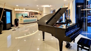 A grand piano in a hotel lobby with a reception desk, framed pictures on the wall, and a modern ceiling design. - De Palma Hotel Shah Alam (Shah Alam)
