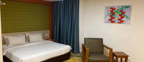 A hotel room with a bed, a chair, a small table, and a painting on the wall.