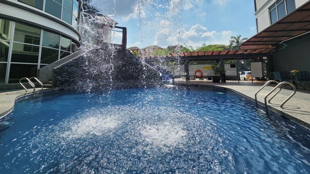 A swimming pool with a water feature, surrounded by buildings with large windows and a covered seating area. - De Palma Hotel Shah Alam (Shah Alam)