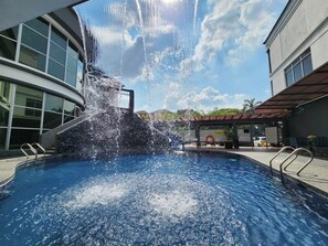 Une piscine agrémentée d’un élément décoratif aquatique, entourée de bâtiments dotés de grandes fenêtres et d’un espace de repos abrité.
