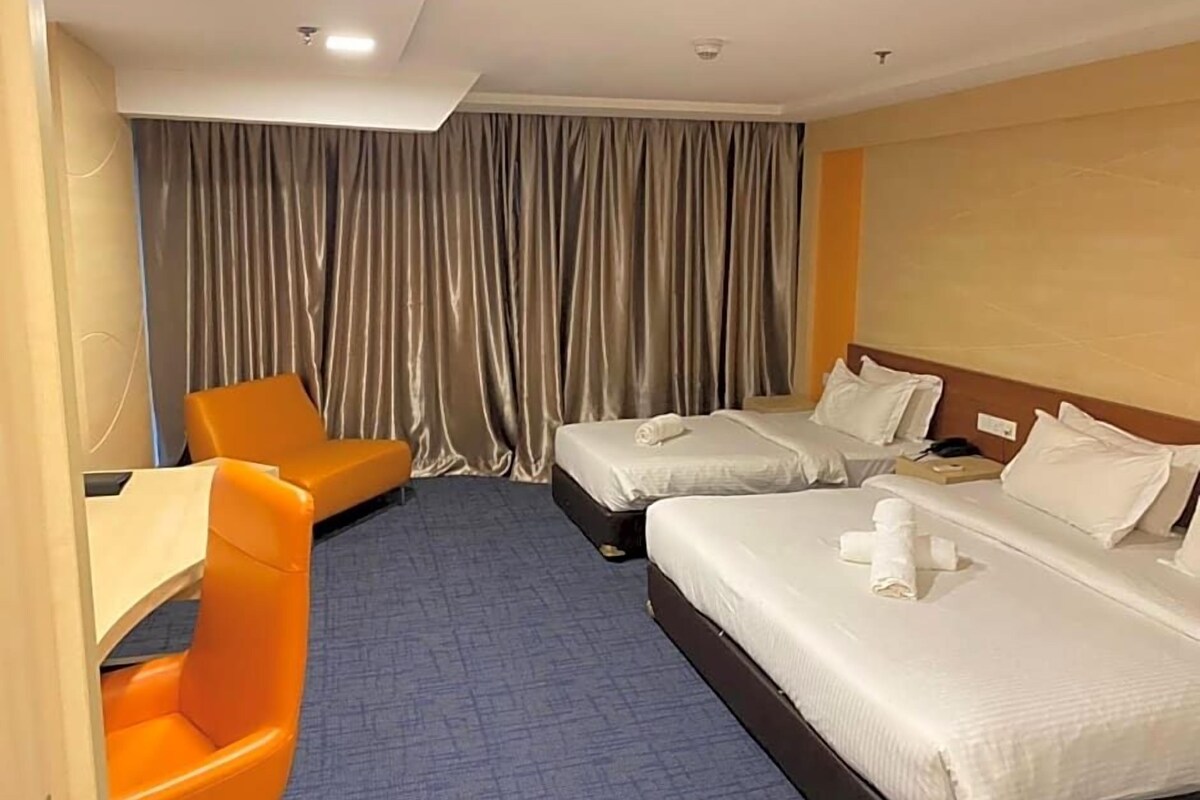 Ein Hotelzimmer mit zwei Betten, einem Schreibtisch, einem orangefarbenen Sessel und einem großen Fenster mit Vorhängen.