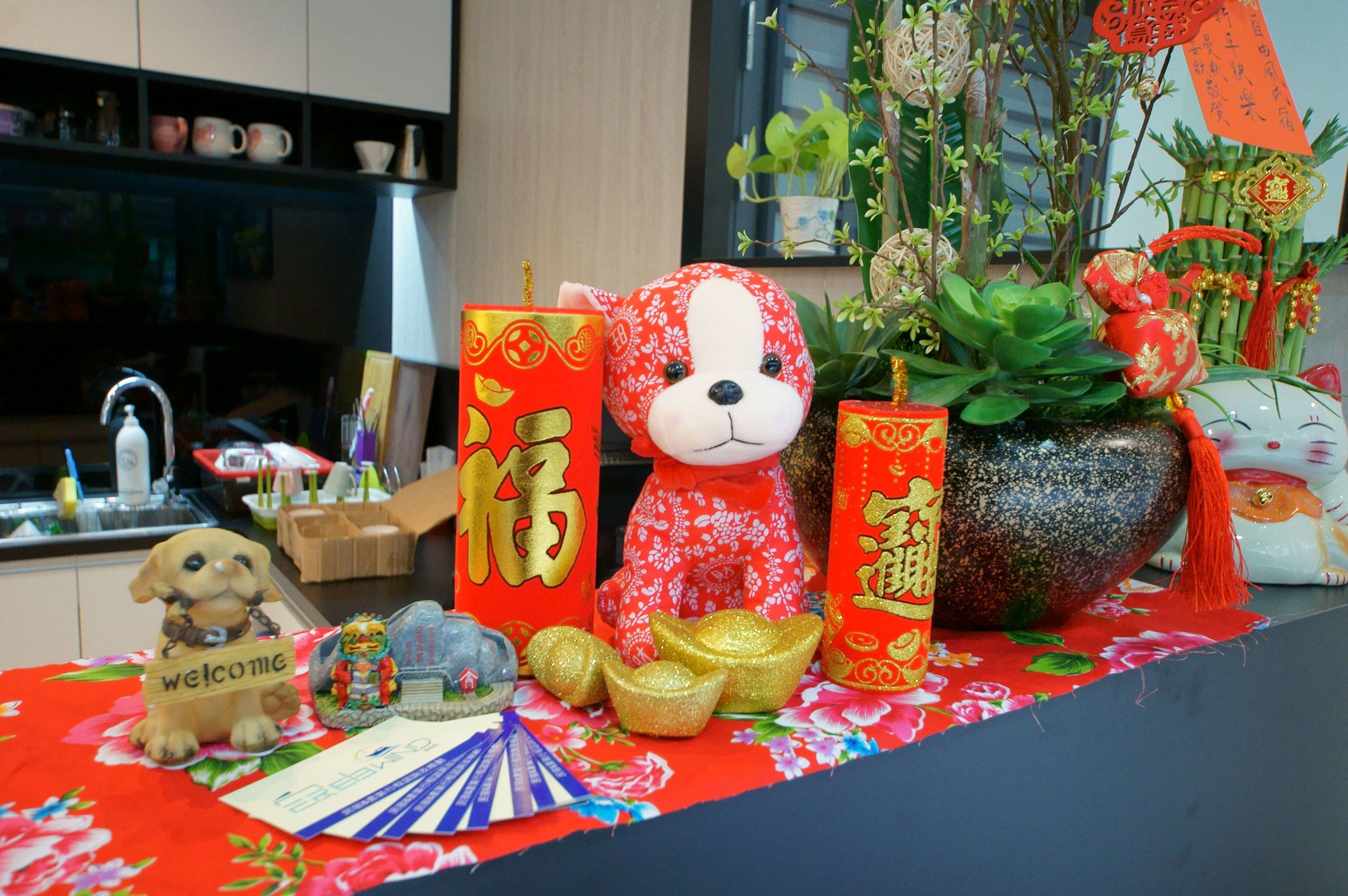A table with a red floral tablecloth, a teddy bear, red and gold decorations, a potted plant, and a small dog figurine.
