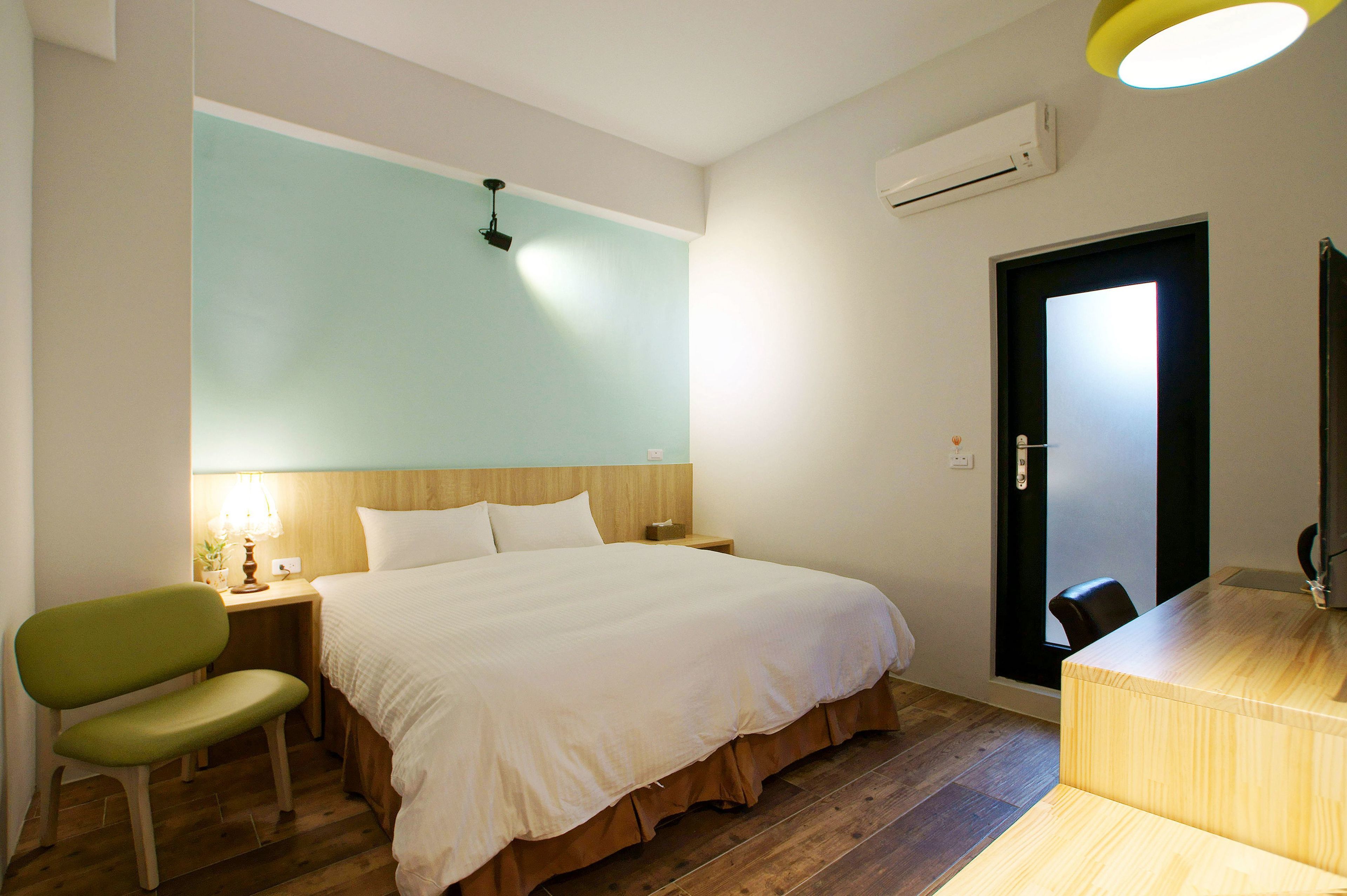 A hotel room with a bed, a desk, a chair, a lamp, and an air conditioning unit.