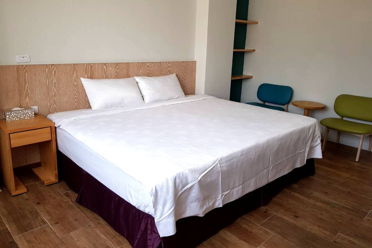 Una cama bien hecha con cabecera de madera, una mesita de noche con caja de pañuelos, y una silla verde en una habitación sencilla con suelo de madera.