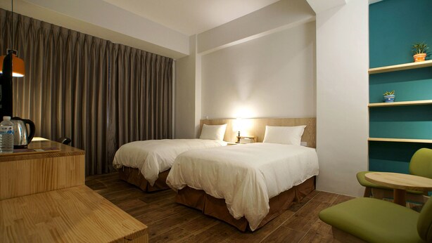 Une chambre d’hôtel avec deux lits, un bureau en bois, une chaise verte et une étagère avec des plantes.