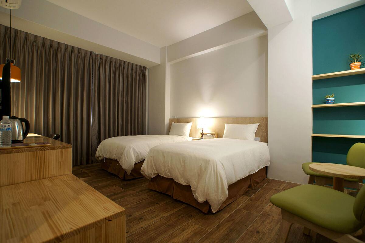 Habitación de hotel con dos camas, un escritorio de madera, una silla verde y una estantería con plantas.