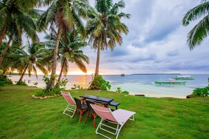 A grassy area with a table and chairs, palm trees, and a view of the sea with boats.