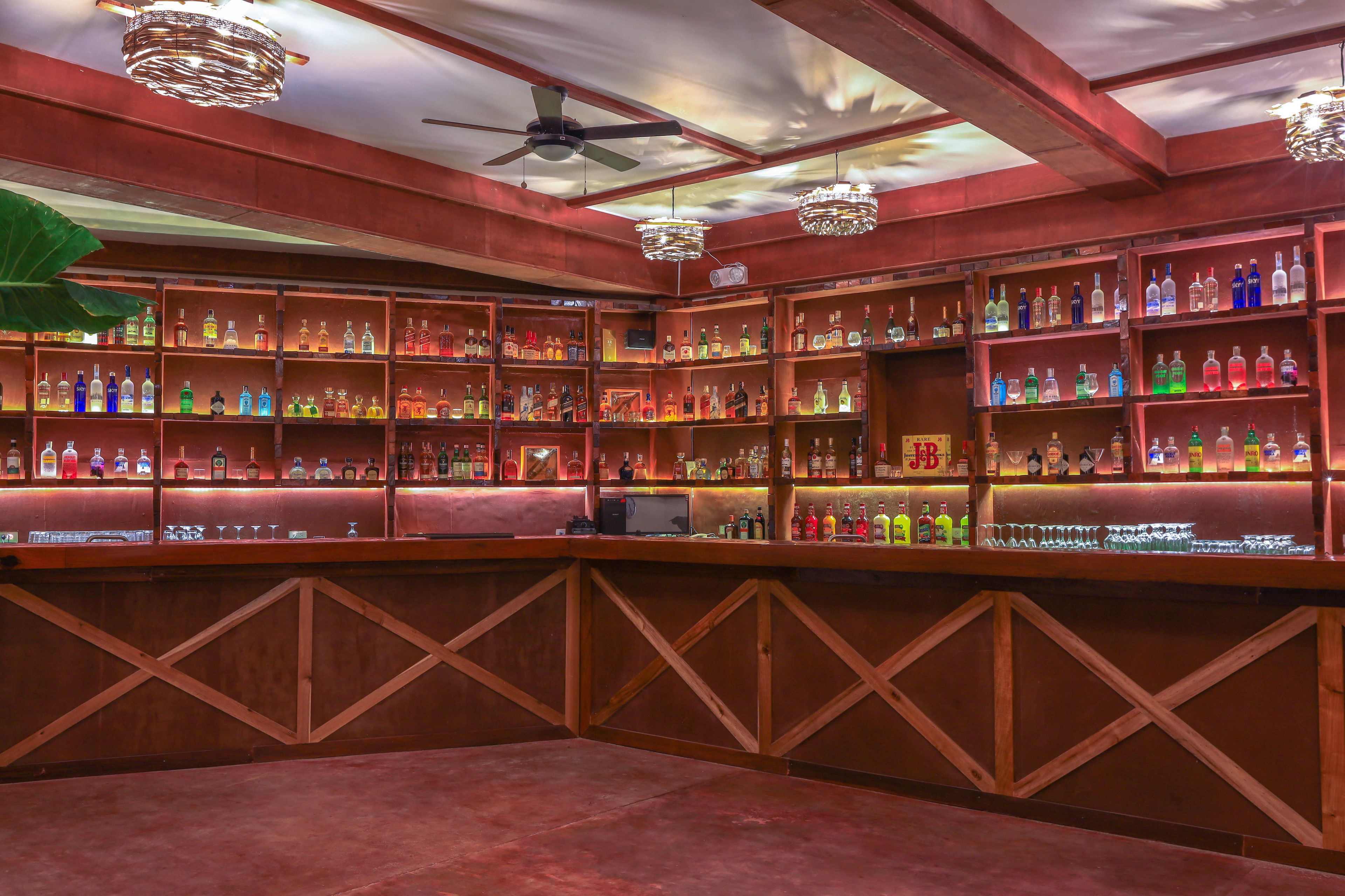 Zona de bar con varias estanterías llenas de botellas de licor, una barra de madera y ventiladores de techo.