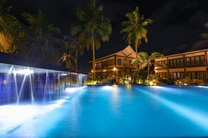 A nighttime view of a hotel with a swimming pool, palm trees, and a waterfall feature.