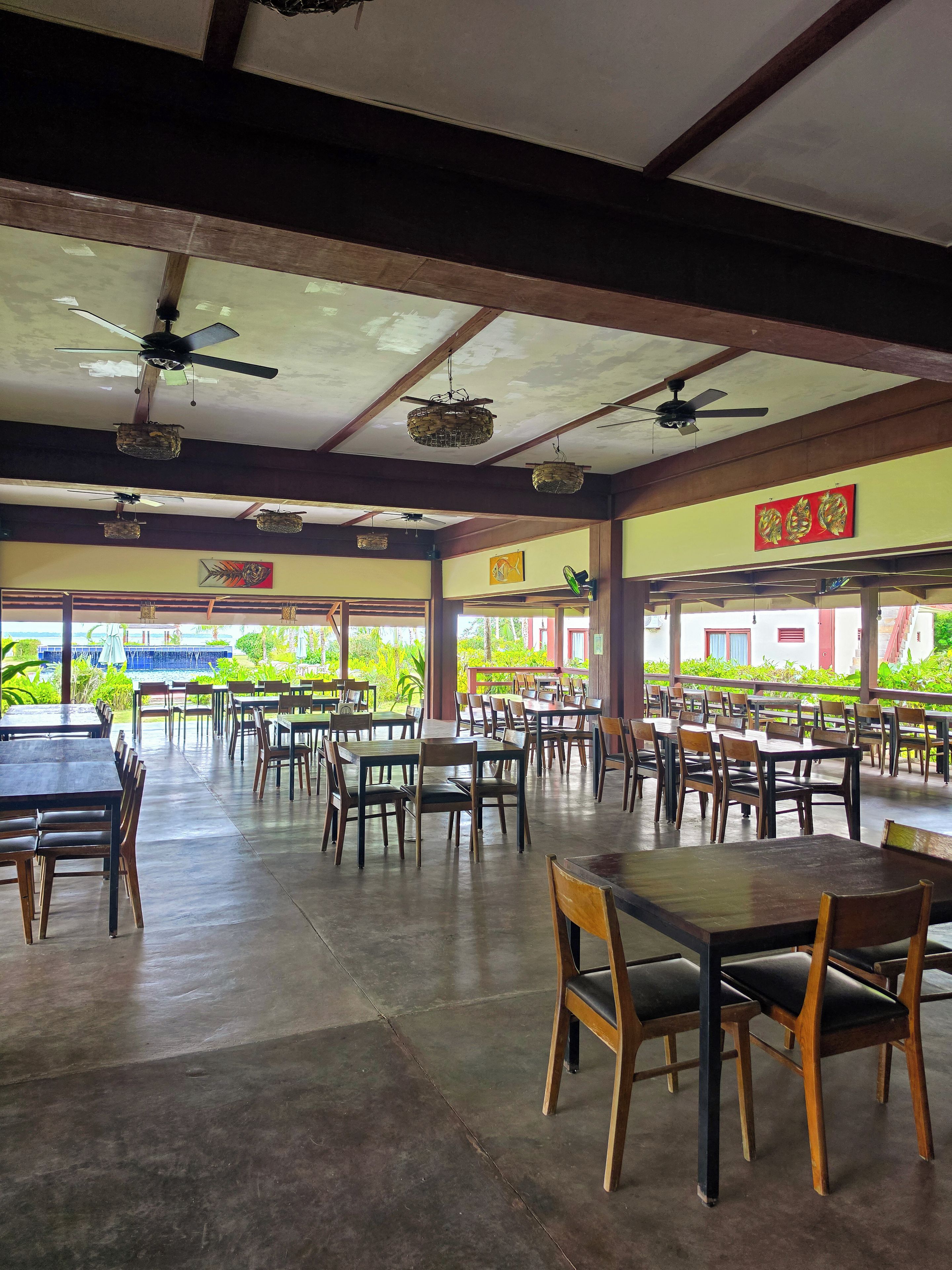 A spacious indoor dining area with multiple tables and chairs, ceiling fans, and a view of greenery outside.