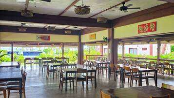 A spacious indoor dining area with multiple tables and chairs, ceiling fans, and a view of greenery outside.
