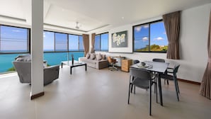 A modern living room with a sofa, dining table, and chairs, large windows with a view of the sea, and a ceiling fan. - Ban Nai Fan (Koh Samui)