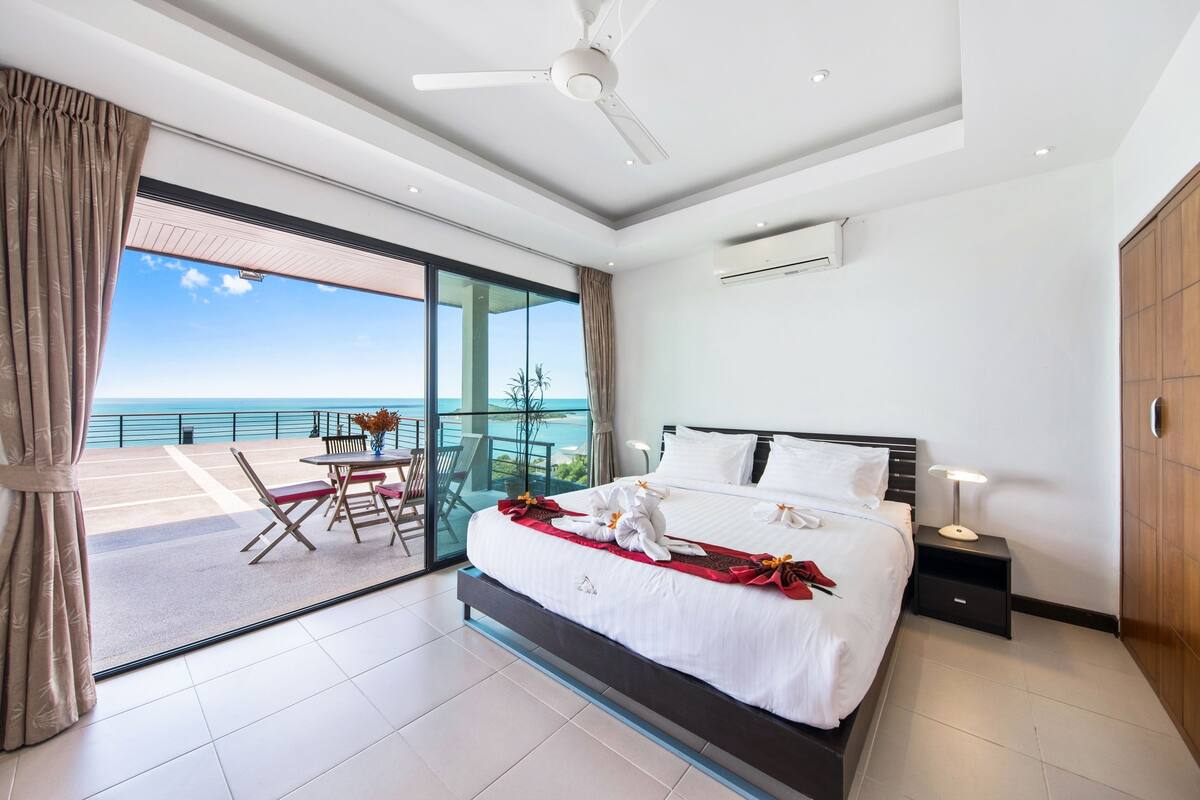 Una habitación de hotel moderna con una cama grande, una mesita de noche, un ventilador de techo y vistas al océano a través de una puerta corredera de vidrio.