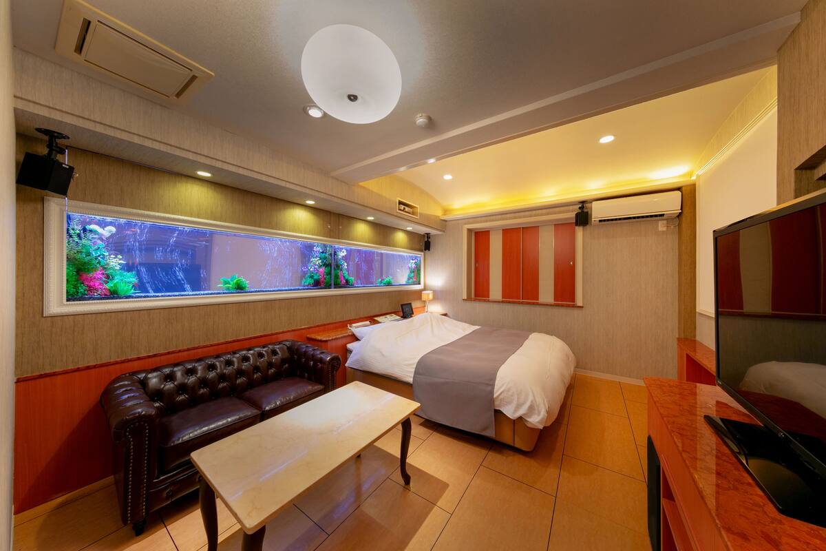 A hotel room with a bed, a leather sofa, a small table, a TV, and a large window with an aquarium.