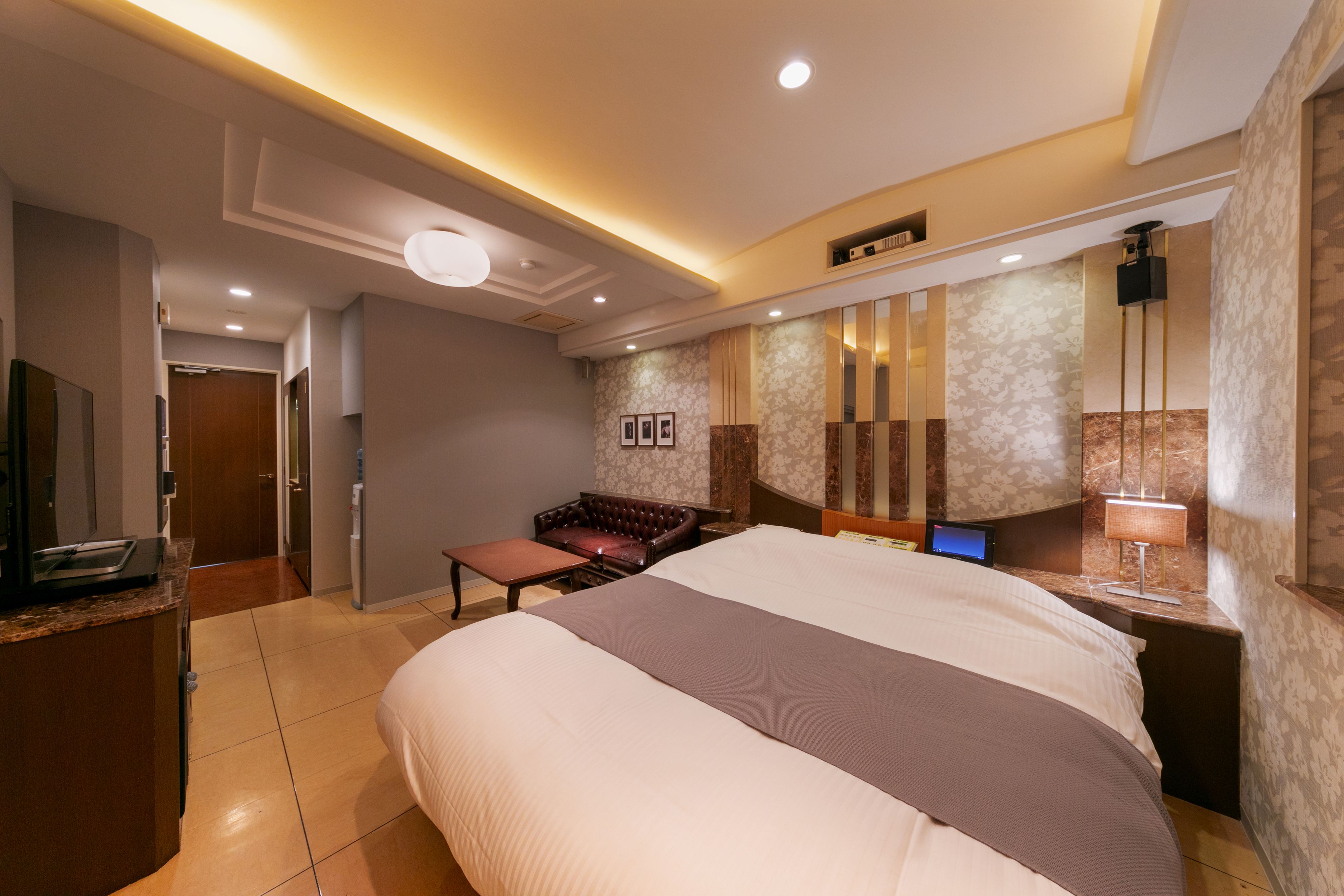 Une chambre d’hôtel dotée d’un grand lit, d’un bureau avec un ordinateur, d’un coin salon avec un canapé et un fauteuil, et d’une salle de bain visible à travers une porte ouverte.