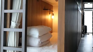 A hotel room with a wooden headboard, a bed with white linens, a wall-mounted light fixture, and a view of the hallway. - Hostel Kura (Tokyo)