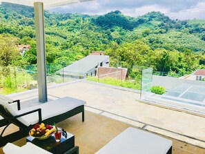 A balcony with a view of a lush green valley and a distant building. There are lounge chairs and a table with fruit and a drink. - Skylight Villa B-01 (Kamala)