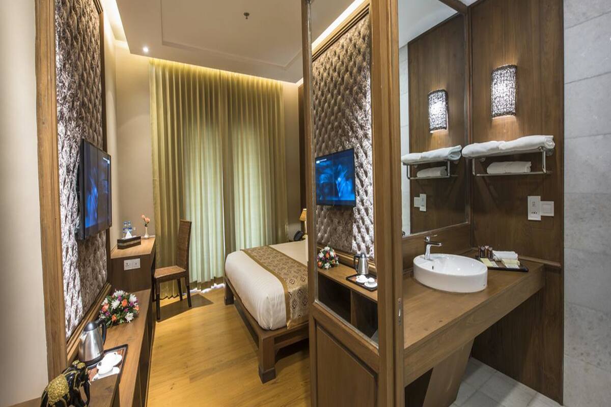Une chambre d’hôtel comprenant un lit, un bureau avec une chaise, une télévision, un grand miroir et une salle de bain avec un lavabo et un porte-serviettes.