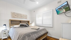 Een slaapkamer met een bed, nachtkastje, lamp, wandtelevisie en een raam met jaloezieën.