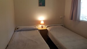A small, dimly lit room with two single beds, a nightstand with a lamp, and a window with curtains.