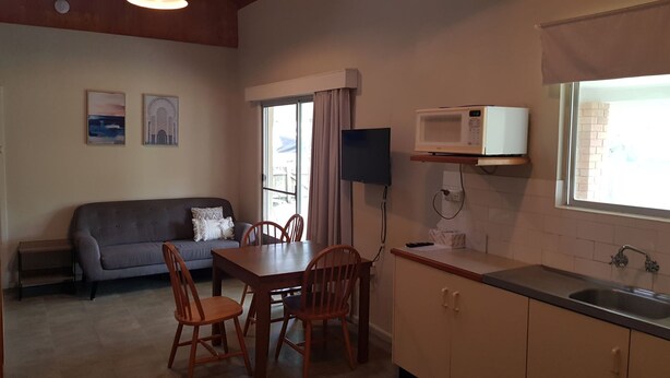 A compact kitchen with a dining table, chairs, a grey sofa, a flat-screen TV, a microwave, and a window with curtains.