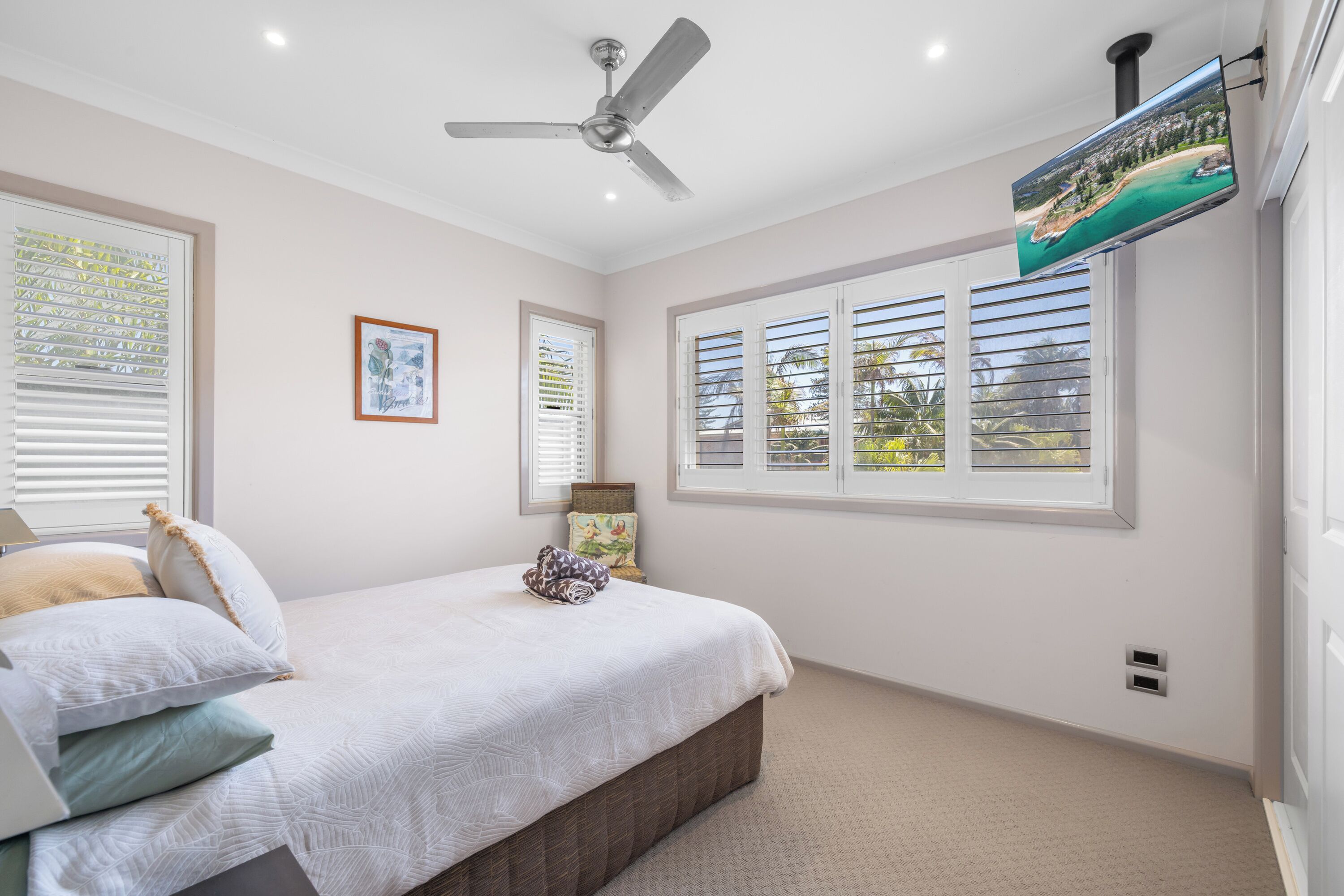 Une chambre à coucher comprenant un lit, un ventilateur de plafond, une fenêtre avec des stores et un téléviseur fixé au mur.