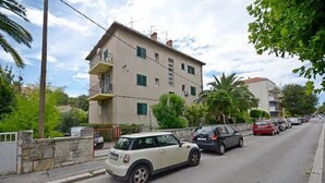 A street scene with a white car parked on the side of the road, a row of parked cars, and residential buildings with balconies and windows. - Elitte Apartment in canter,self check in /checkout (Split)