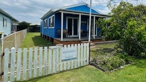 Une maison mobile bleue, dotée d’une véranda, entourée d’une clôture blanche en bois et située sous un ciel partiellement nuageux.