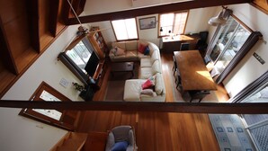 A high-ceilinged living room with a wooden staircase, a sofa set, a dining table, and a view through a large window. - Kookaburra Cottage 71 Straight Hat Head (Hat Head)