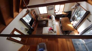 A high-ceilinged living room with a wooden staircase, a sofa set, a dining table, and a view through a large window.