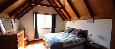 Une chambre avec un plafond en pente, un lit recouvert d’une courtepointe, une commode, une table de chevet avec une lampe et une fenêtre avec des rideaux.