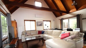 A spacious living room with a high ceiling, large windows, and a wooden ceiling beam. - Kookaburra Cottage 71 Straight Hat Head (Hat Head)