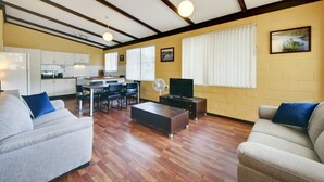 Ein Wohnzimmer mit Sofa, Couchtisch und Fernseher. Im Hintergrund ist eine Küche mit Schränken und Geräten zu sehen.