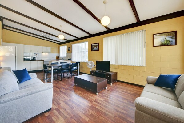 A living room with a sofa, coffee table, and TV. There is a kitchen in the background with cabinets and appliances.