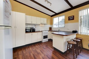 Une cuisine avec des meubles blancs, un four à micro-ondes, une cuisinière et un évier. On y trouve des tabourets en bois et une fenêtre avec des stores.