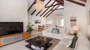 A spacious living room with a large flat-screen TV, a wooden coffee table, a white sofa, and a dining area in the background.
