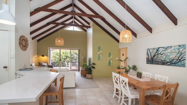 A spacious kitchen with a high ceiling, wooden beams, a dining area with white chairs, and a large window providing natural light.
