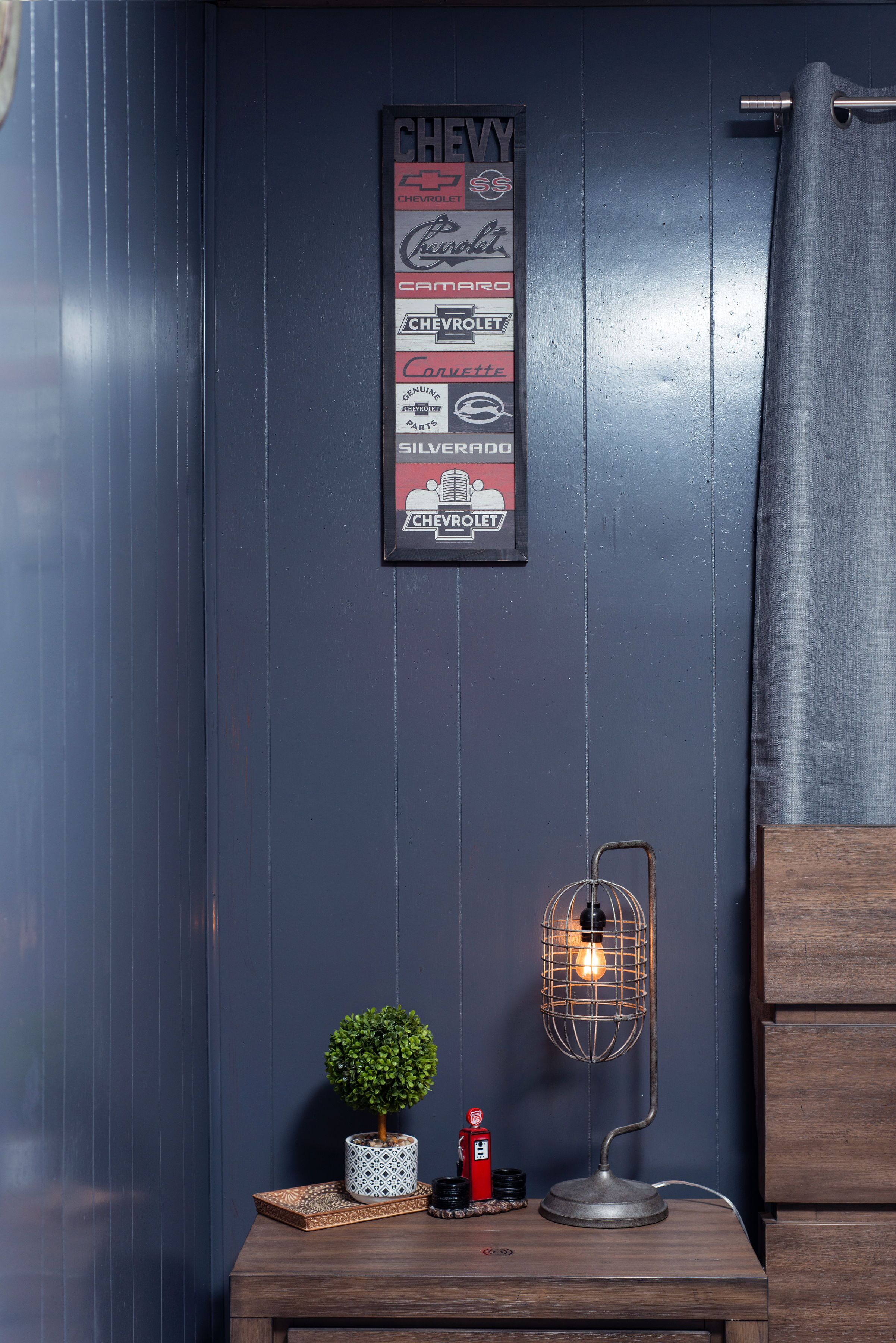 A wall with a Chevrolet-themed sign, a small potted plant, a vintage-style lamp, and a red fire extinguisher on a wooden table.
