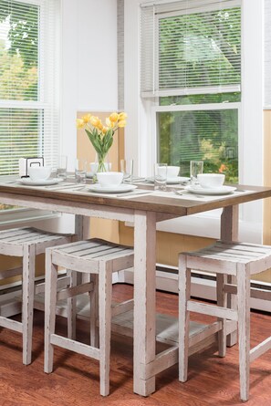 Une table à manger est dressée avec de l’assiette, des bols et des verres blancs, et est entourée de bancs en bois blancs, dans une pièce dotée de grandes fenêtres et offrant une vue sur la verdure.