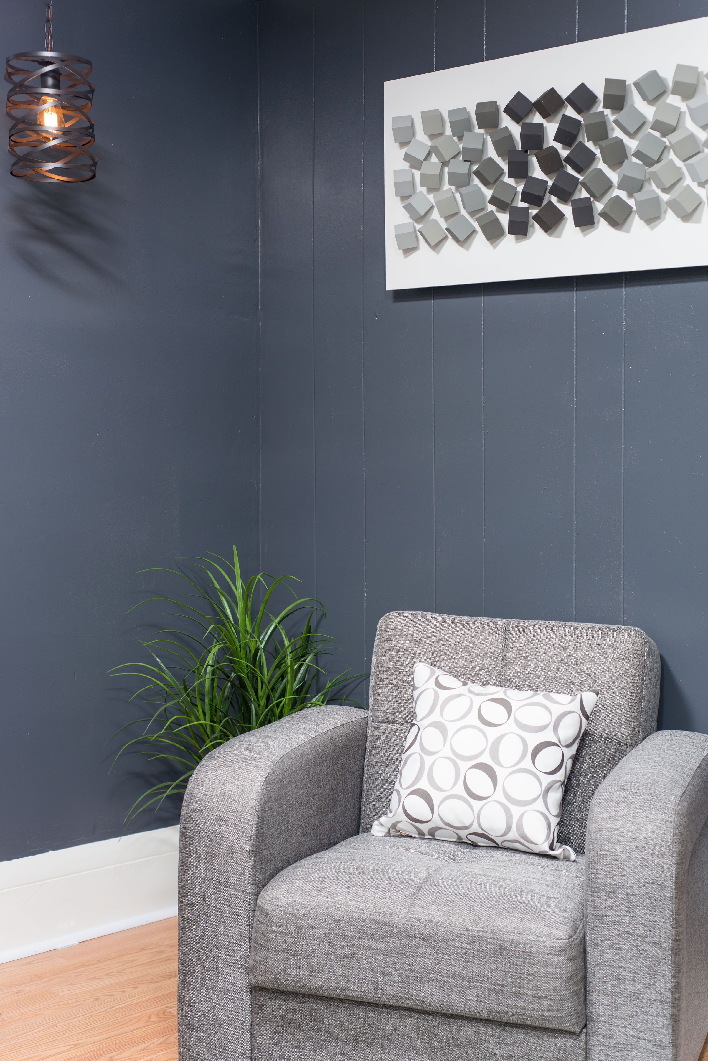 A grey armchair with a patterned pillow against a dark wall, a hanging light fixture, and a plant.